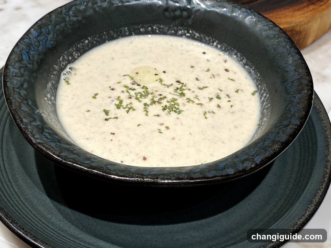 A bowl of creamy white soup, likely a bisque or chowder, garnished with chopped green herbs and a drizzle of oil. The soup is served in a dark, textured ceramic bowl placed on a matching saucer.