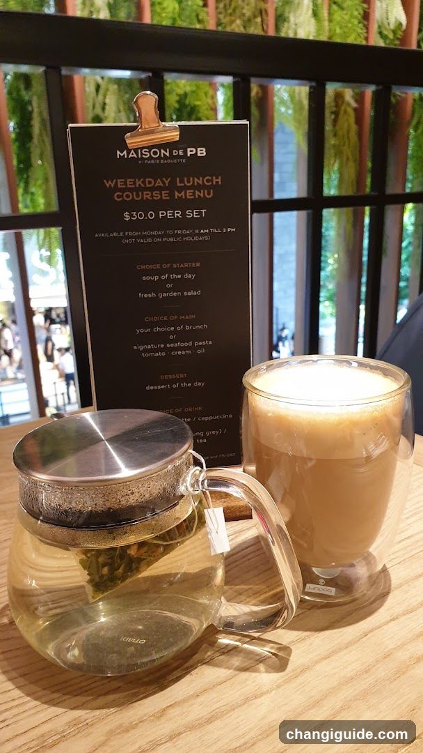 A table setting featuring a glass tea infuser with green leaves, a frothy coffee drink in a double-walled glass, and a menu board titled 'MAISON DE PB - PARIS BARISTE WEEKDAY LUNCH COURSE MENU $30.0 PER SET'. The background shows an outdoor seating area with hanging plants.