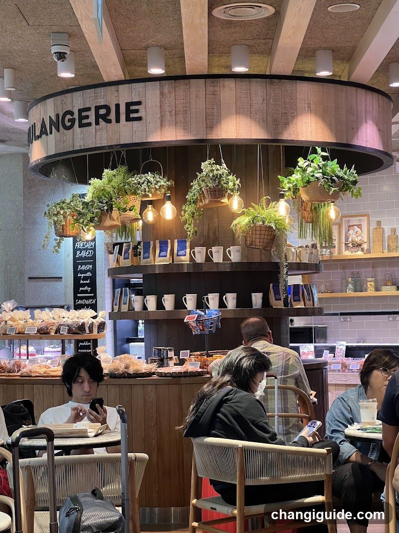 Interior of a boulangerie with customers seated at tables, hanging plants, and shelves displaying coffee mugs and packaged goods.