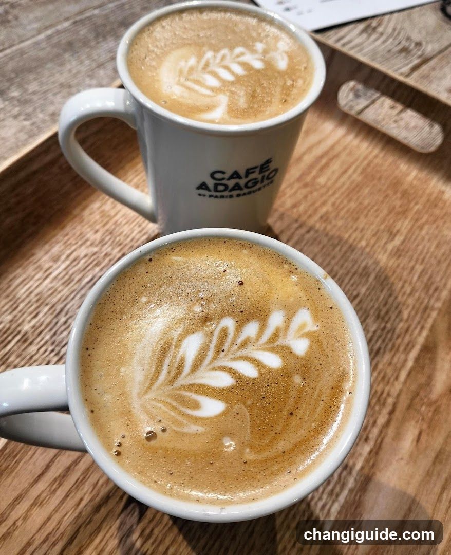 Two white ceramic mugs filled with coffee featuring leaf-shaped latte art, placed on a wooden tray. The mug in the background has 'CAFÉ ADAGIO' printed on it.