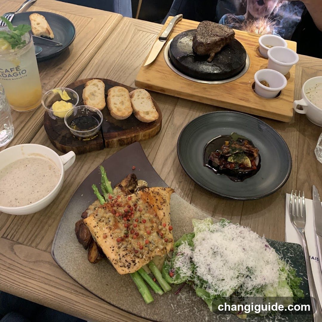 A table at Cafe Baglio featuring a variety of dishes including steak on a hot stone, bread slices, asparagus, salad with parmesan cheese, and drinks in branded cups.
