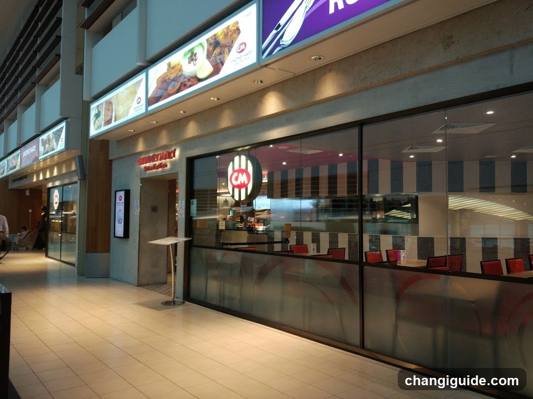 A modern restaurant storefront with large glass windows and a visible entrance. The CM logo is prominently displayed on the window, along with illuminated signage above. Inside, red chairs and tables are arranged neatly.
