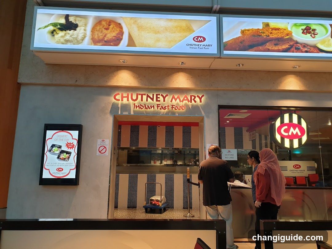 The image shows the storefront of Chutney Mary, an Indian fast food outlet. The shop has a sign with the name 'CHUTNEY MARY' and 'Indian Fast Food' written in illuminated letters above the entrance. There are two large digital screens displaying images of food items. A person is standing near the counter inside the shop, while another individual appears to be waiting outside.