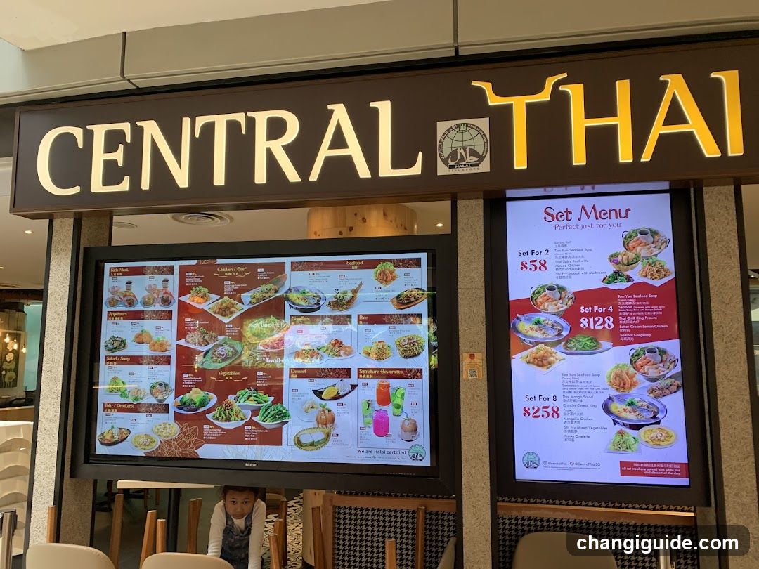 The image shows the storefront of a restaurant named 'CENTRAL THAI'. The sign is prominently displayed above the entrance with illuminated lettering and a logo featuring a globe. Below the sign, there are two large digital screens displaying menus. One screen showcases various dishes with images and descriptions, while the other highlights set menu options with prices. A person is partially visible sitting inside the restaurant in the lower left corner.
