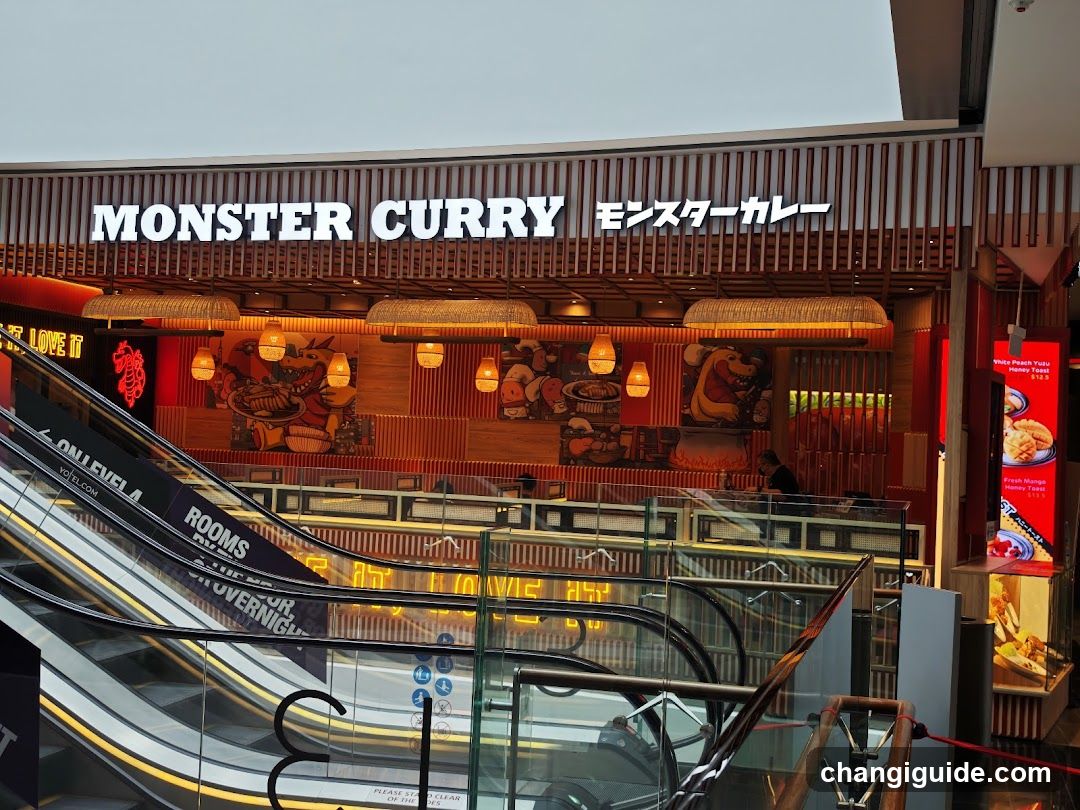 The photo shows the storefront of Monster Curry, a restaurant chain with Japanese and Western fusion cuisine. The sign above reads 'MONSTER CURRY' in English and Japanese characters. Below the sign are decorative elements including hanging lights and artwork depicting curry dishes. An escalator is visible in front of the store.