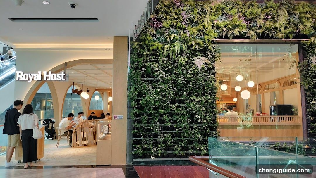 The image shows the storefront of a shop named 'Royal Host', with visible signage on the building facade. The entrance features an arched design, and there is a lush green vertical garden wall to the right. Inside, customers are seated at tables, and warm lighting hangs from the ceiling.