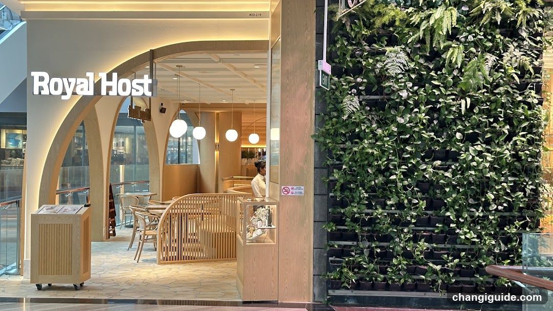 The photo shows the storefront of a shop named 'Royal Host', with its signage clearly visible on the building facade. The entrance features an arched design, wooden furniture inside, and hanging spherical lights. A lush green vertical garden is adjacent to the shop.