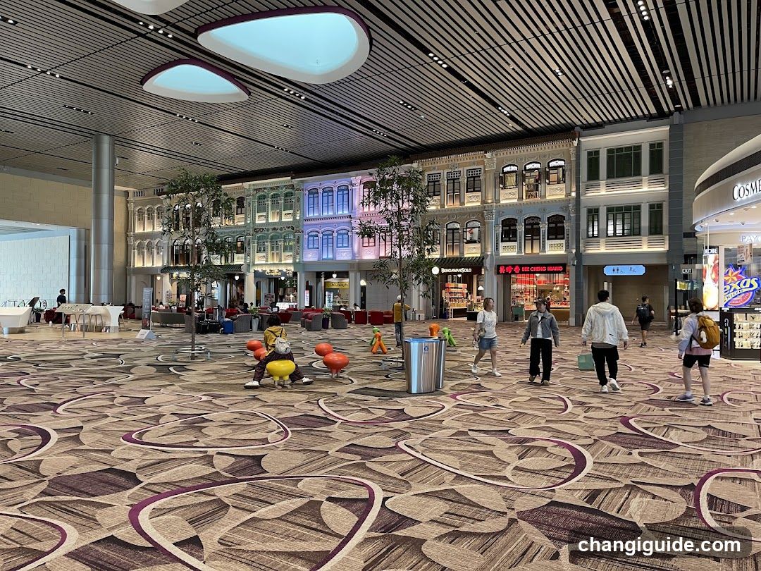 A spacious airport terminal with patterned carpet, decorative ceiling lights, and a row of shops with illuminated facades in the background. People are walking or sitting on colorful benches.