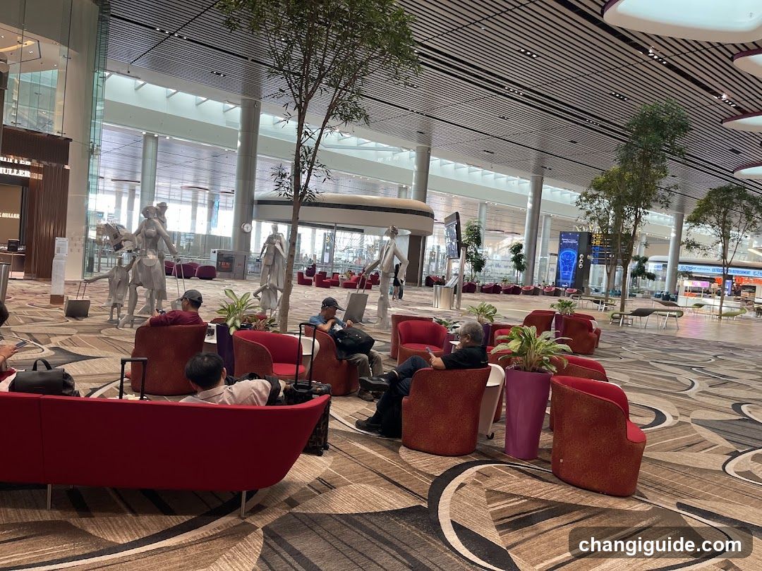 A spacious airport lounge with red seating, patterned carpeting, and large windows overlooking the terminal. Several people are seated or walking, some with luggage. Decorative statues and potted plants add to the ambiance.