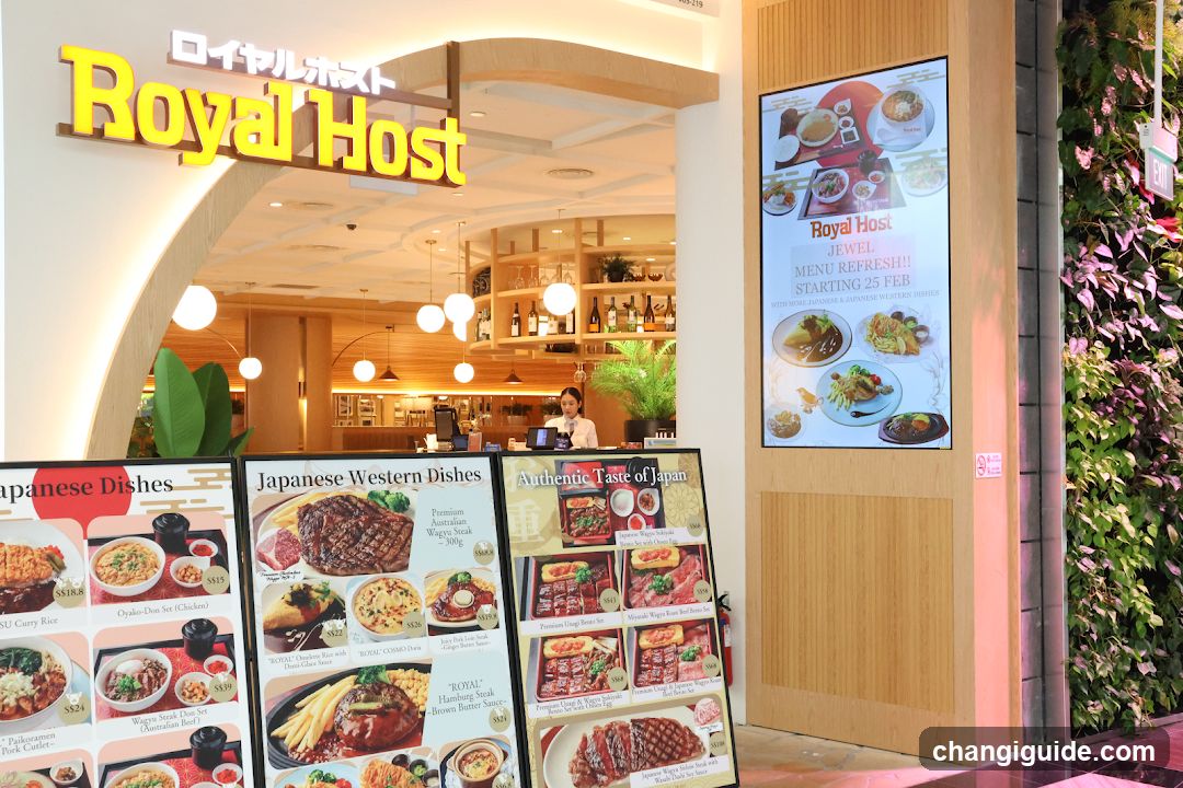 Exterior view of Royal Host restaurant entrance with illuminated signage, wooden facade, and vertical garden wall. Menu boards display Japanese dishes and western cuisine options.