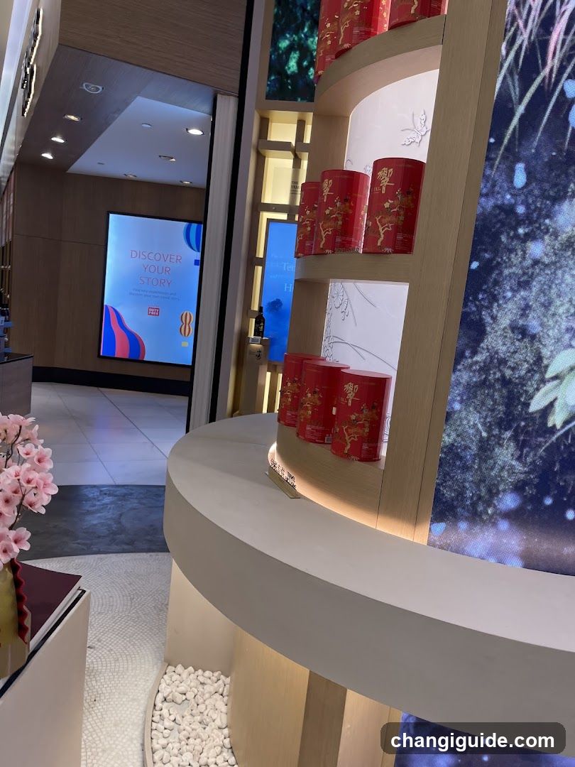 Interior view of a tea shop featuring curved wooden shelves displaying red cylindrical tea tins with gold lettering. A digital screen in the background shows 'DISCOVER YOUR STORY' alongside colorful abstract graphics and a logo resembling a stylized teapot or flame. Pink flowers are visible on the left, white pebbles line the floor near the base of the display, and ambient lighting highlights the merchandise.