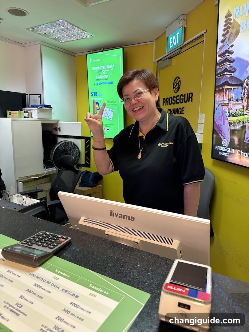 A smiling staff member in a black Prosecur Change uniform stands behind the counter, making a peace sign gesture. The background features yellow walls with signage including an exit sign and posters advertising travel packages to Bali. On the desk there is a calculator, a payment terminal, and printed price lists showing data plans like Unlimited SG Data at $18.