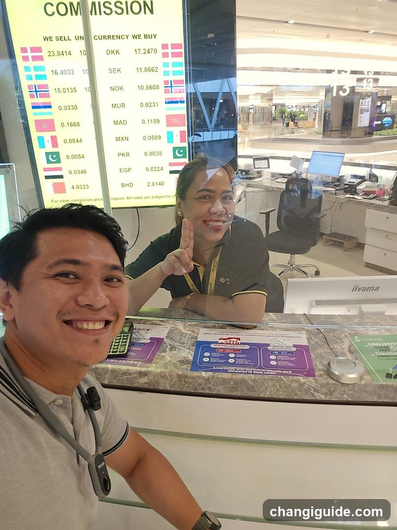 A man and a woman are smiling at the camera in what appears to be an airport or mall currency exchange booth. The woman is behind the counter, wearing a uniform with a lanyard, and making a peace sign. Behind her is a large digital display showing exchange rates for various currencies like DKK, SEK, NOK, MAD, MXN, PKR, EGP, BHD. There are informational brochures on the counter surface.