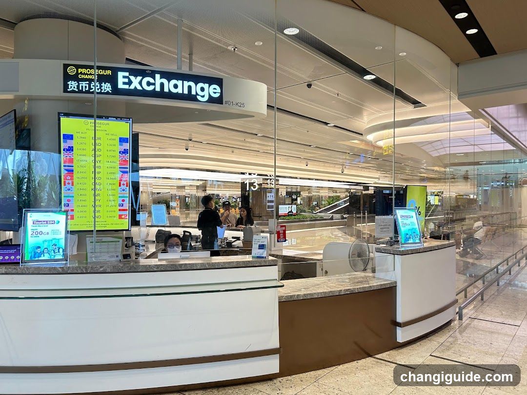A currency exchange counter with a glass facade and digital display showing various currency rates. Staff members are visible behind the counter, and promotional materials are placed on the counter surface.