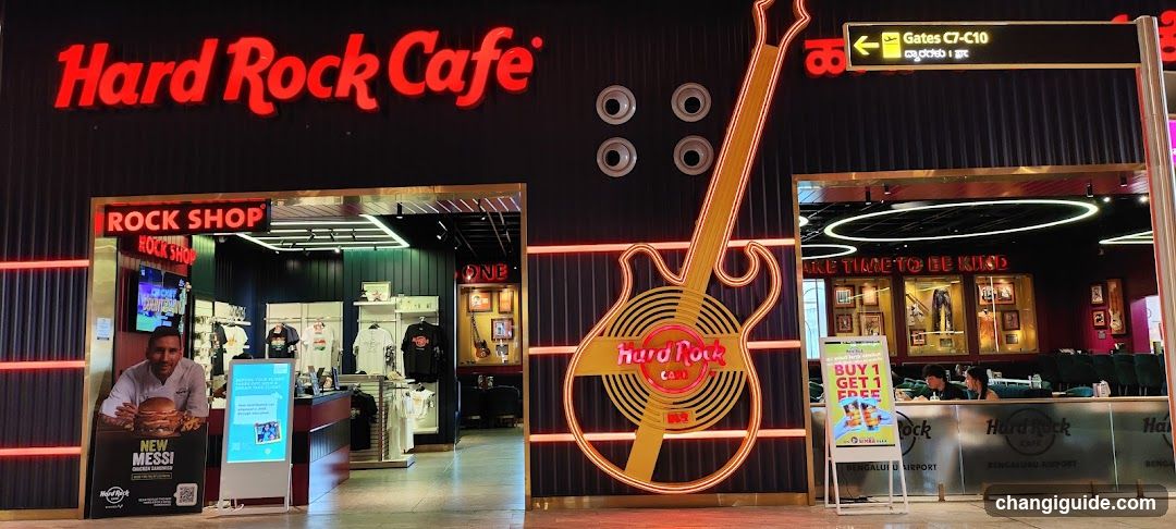 The image shows the exterior facade of a Hard Rock Cafe with an adjacent Rock Shop. The building features illuminated signage including 'Hard Rock Cafe' in red neon lettering above the entrance. A large neon guitar graphic is mounted on the wall between two glass doors leading into separate areas labeled 'ROCK SHOP' and another section displaying merchandise. Inside, racks of clothing are visible along with promotional posters and displays. A sign indicates airport gates C7-C10 nearby.
