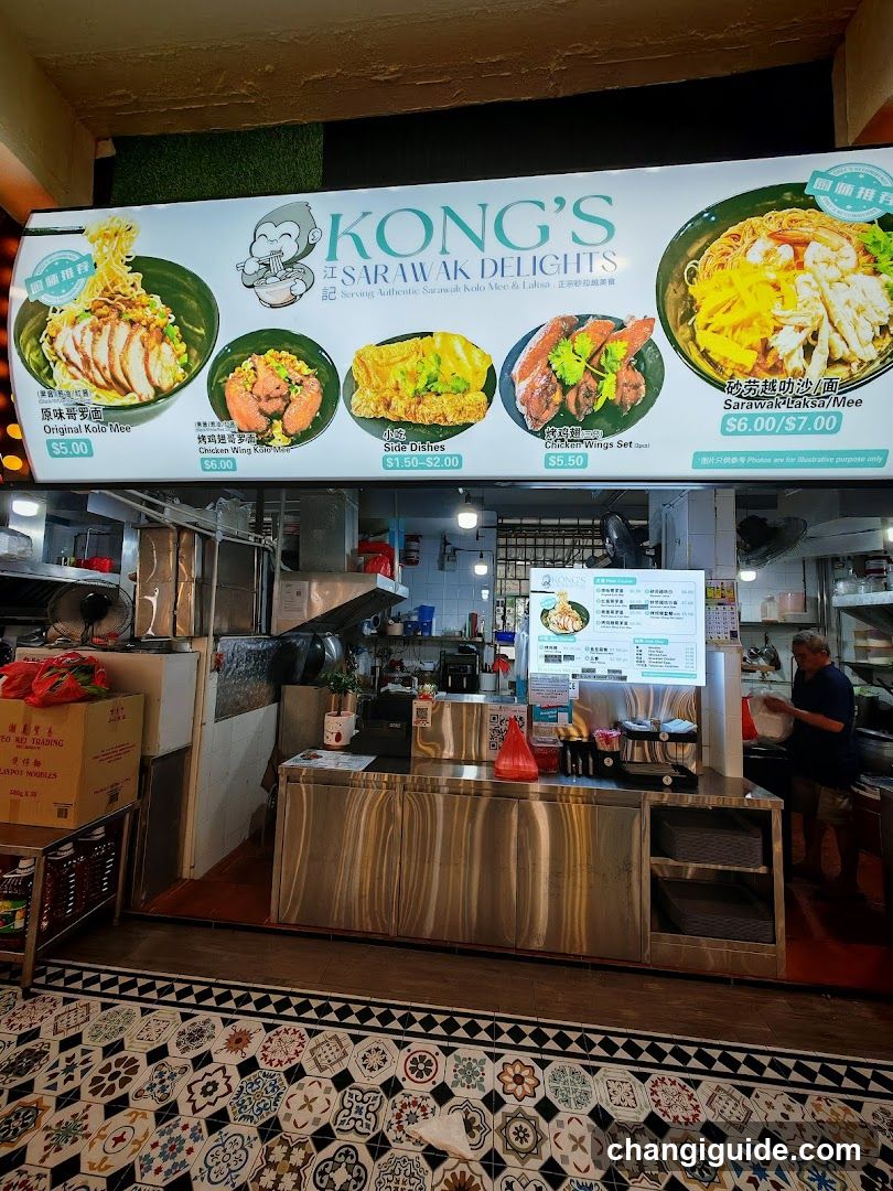 A food stall with a large illuminated menu board displaying various dishes like Original Yum Mee, Chicken Wings, Side Dishes, and Saraman Chicken Rice. The interior shows stainless steel counters, cooking equipment, and staff preparing food.